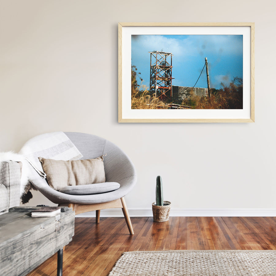 A rusting steel lattice headframe with two spoked sheave wheels silhouetted against a bright sky at Ikeshima Coal Mine, with dry grasses blurring across the foreground.