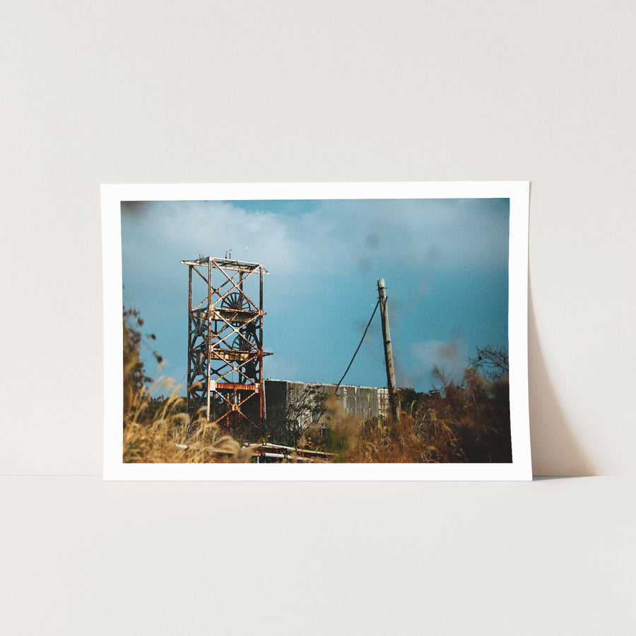 A rusting steel lattice headframe with two spoked sheave wheels silhouetted against a bright sky at Ikeshima Coal Mine, with dry grasses blurring across the foreground.