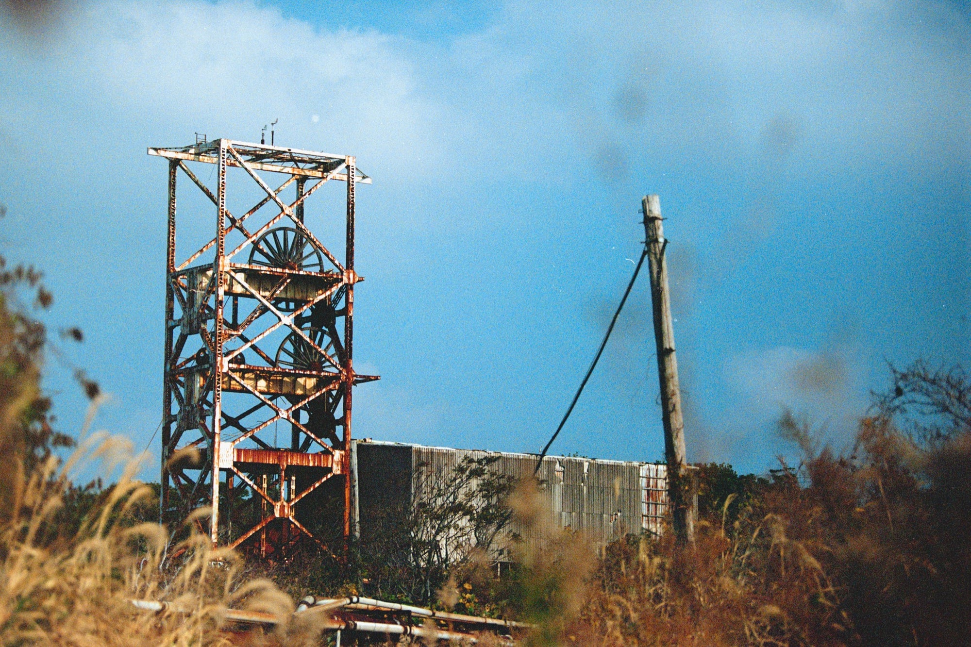 A rusting steel lattice headframe with two spoked sheave wheels silhouetted against a bright sky at Ikeshima Coal Mine, with dry grasses blurring across the foreground.