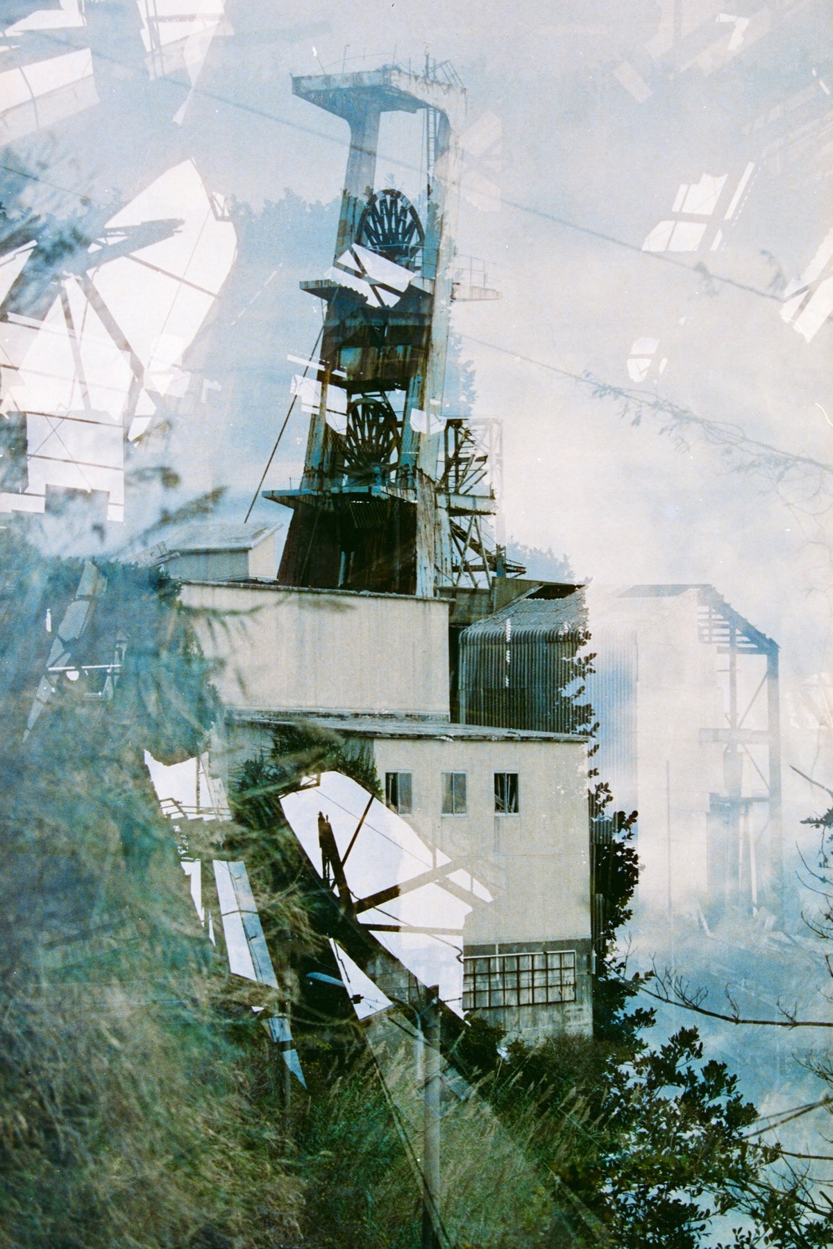 Multiple exposures stack the steel headframe and spoked winding wheels of Ikeshima Coal Mine above a rendered engine house, with blurred foliage across the foreground and a corrugated metal structure to the right.