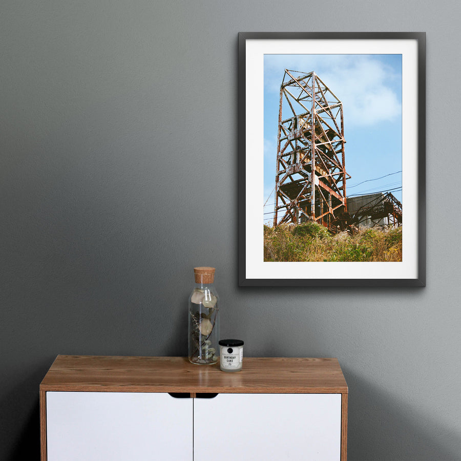 Rusted steel lattice of a mine headframe at Ikeshima, with power lines crossing toward a corrugated shed and dry golden grass on the slope below.