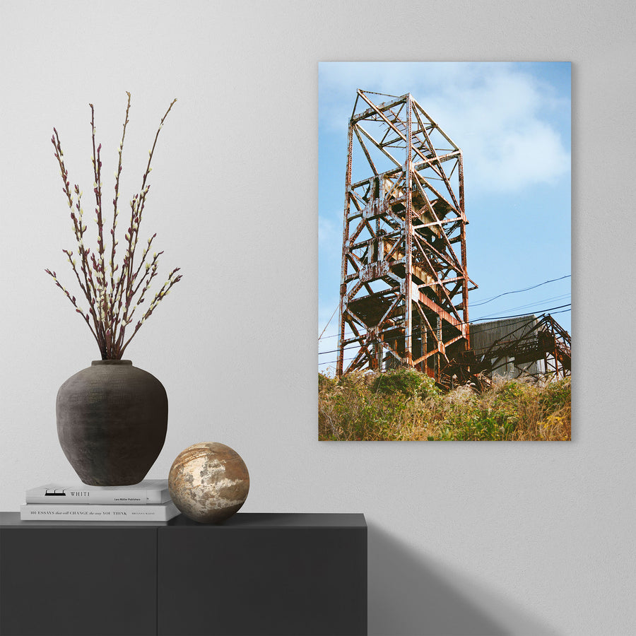 Rusted steel lattice of a mine headframe at Ikeshima, with power lines crossing toward a corrugated shed and dry golden grass on the slope below.
