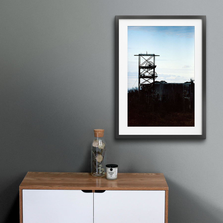 The twin spoked winding wheels of the steel lattice headframe at Ikeshima Coal Mine, silhouetted against a warm sunset sky, with dense foliage surrounding the lower framework and a corrugated metal building visible to the right.