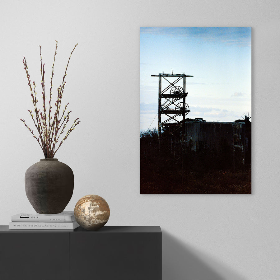 The twin spoked winding wheels of the steel lattice headframe at Ikeshima Coal Mine, silhouetted against a warm sunset sky, with dense foliage surrounding the lower framework and a corrugated metal building visible to the right.