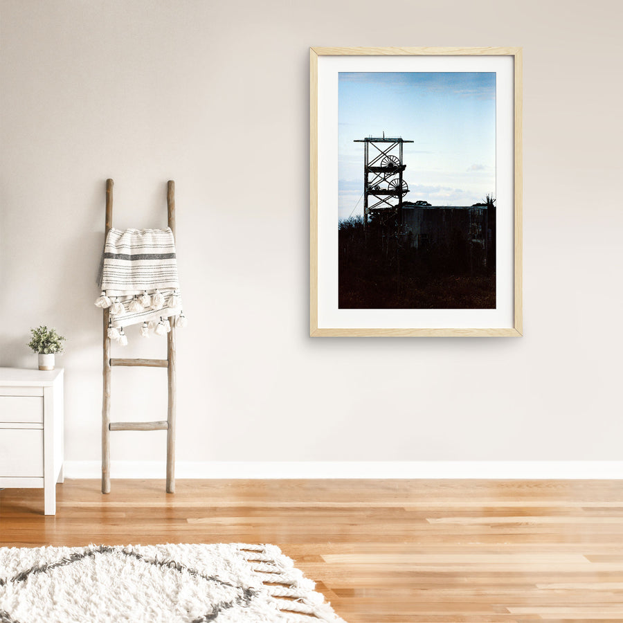 The twin spoked winding wheels of the steel lattice headframe at Ikeshima Coal Mine, silhouetted against a warm sunset sky, with dense foliage surrounding the lower framework and a corrugated metal building visible to the right.
