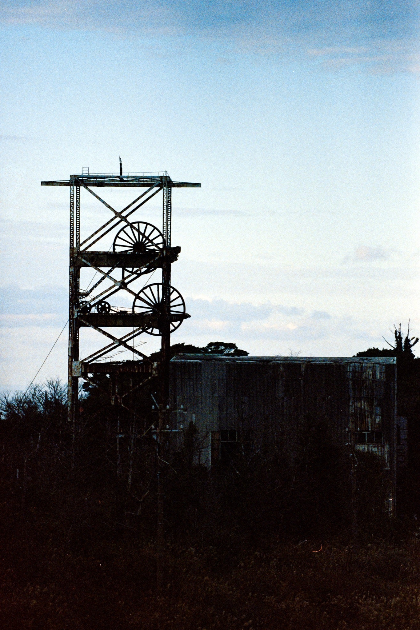 The twin spoked winding wheels of the steel lattice headframe at Ikeshima Coal Mine, silhouetted against a warm sunset sky, with dense foliage surrounding the lower framework and a corrugated metal building visible to the right.