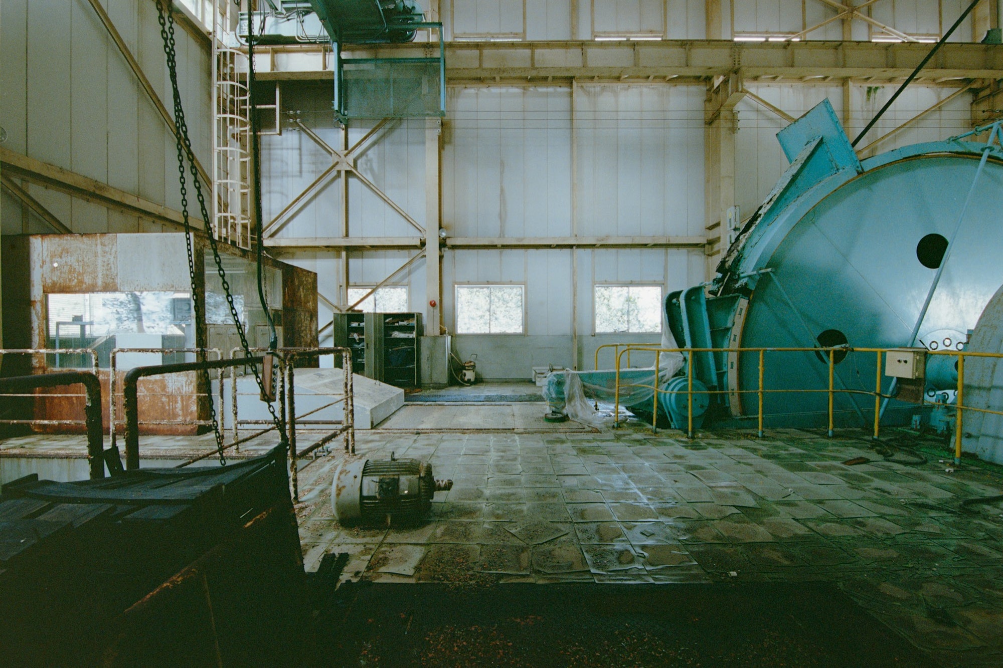 The sheave wheel, hoist drum and hanging chains of the winding house interior at Ikeshima Coal Mine, with a yellow-railed cylindrical vessel to the right and three rectangular windows along the far wall.