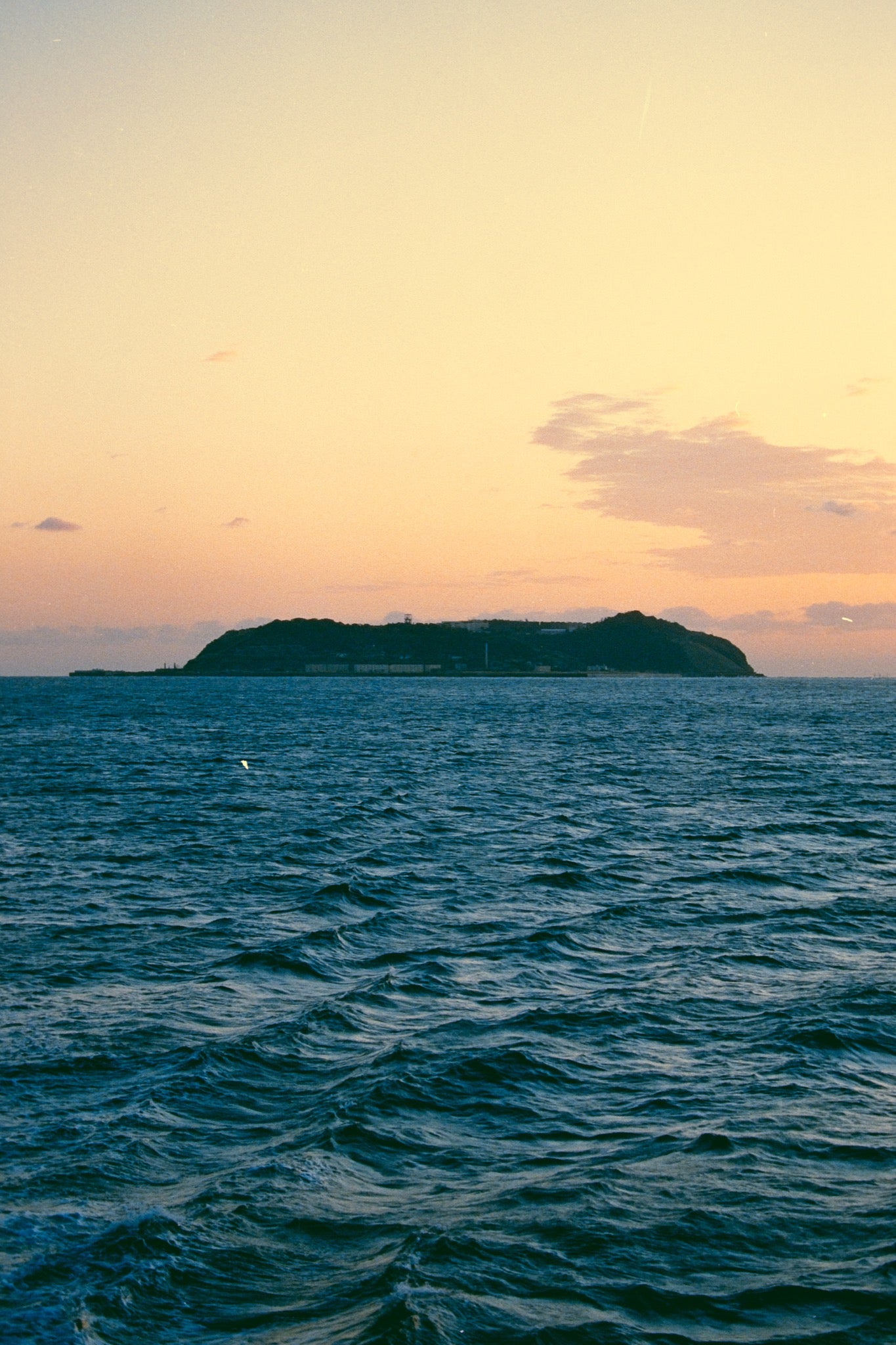 Ikeshima seen as a low dark silhouette across the Gotō Sea at dusk, a contrail crossing a peach-and-yellow sunset sky above.
