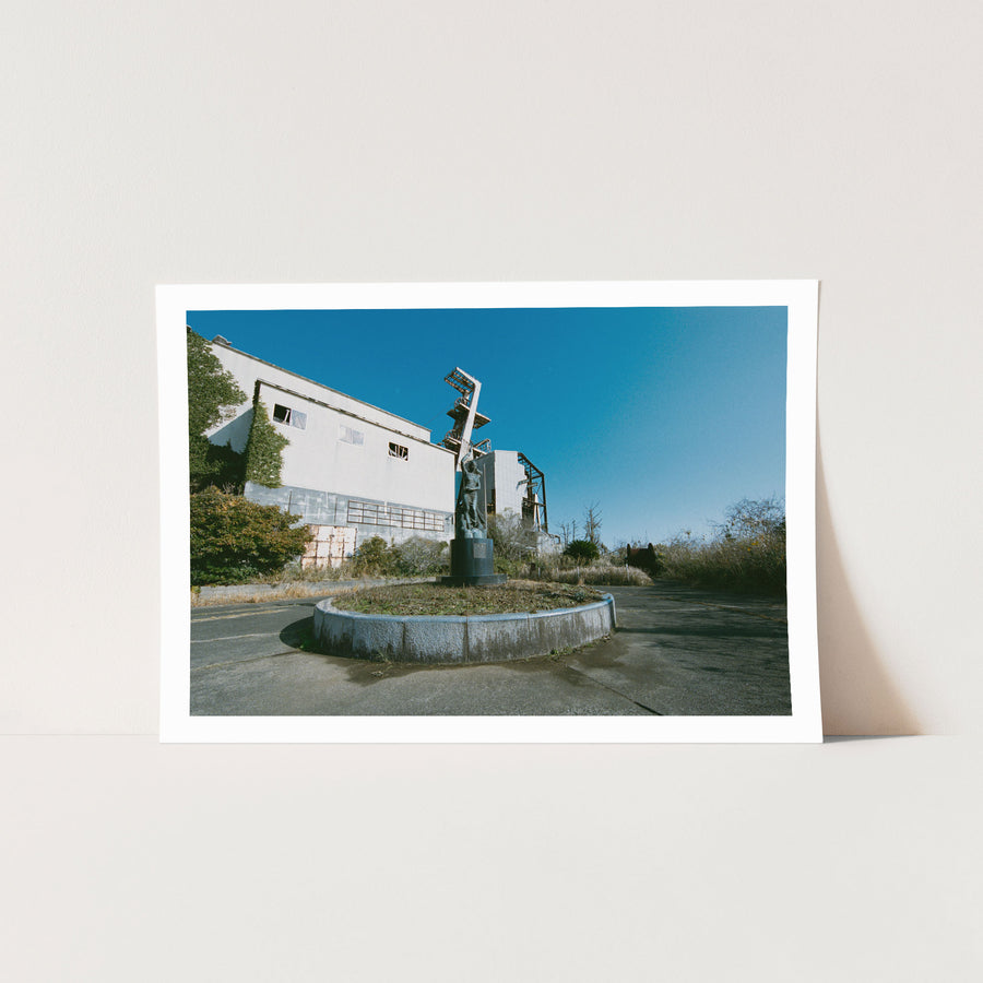 A weathered bronze statue of a woman with arms raised on a cylindrical plinth, surrounded by dry grass and scrub, with a rusted abandoned building and the Ikeshima Coal Mine winding house and headframe behind her.