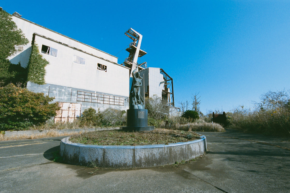 A weathered bronze statue of a woman with arms raised on a cylindrical plinth, surrounded by dry grass and scrub, with a rusted abandoned building and the Ikeshima Coal Mine winding house and headframe behind her.