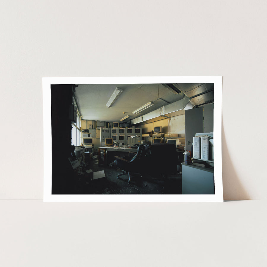 A bank of twelve dark CRT monitors set into and stacked above a central control console at the Ikeshima Coal Mine, with monthly report binders on a shelf to the right and daylight entering through a tall window at left.