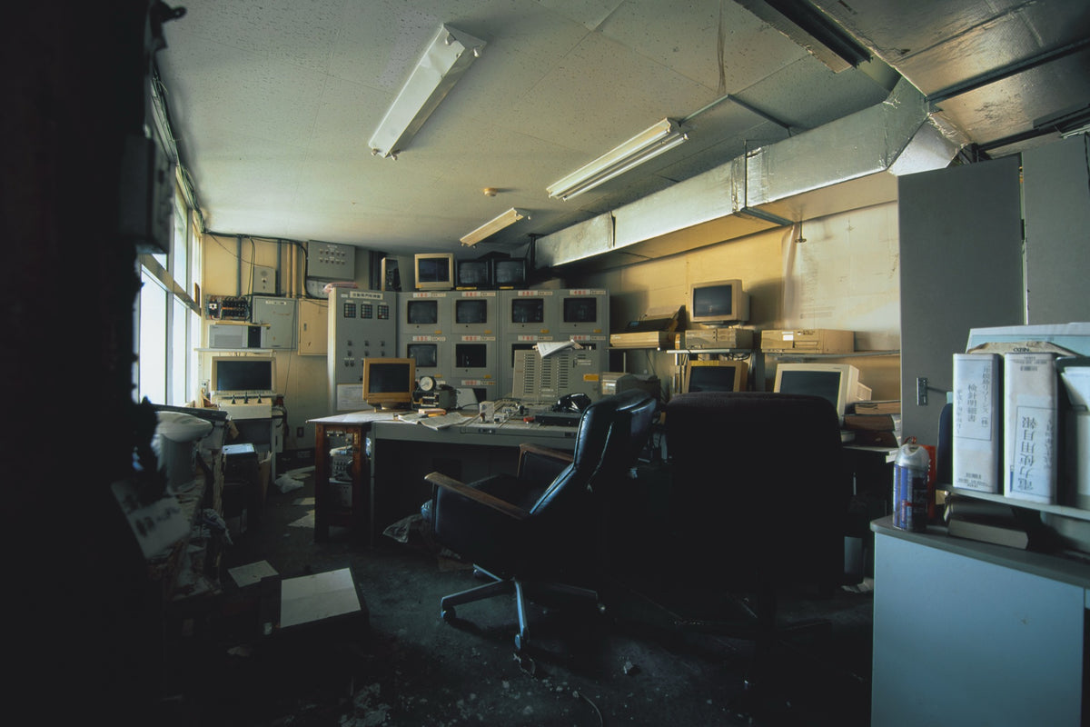 A bank of twelve dark CRT monitors set into and stacked above a central control console at the Ikeshima Coal Mine, with monthly report binders on a shelf to the right and daylight entering through a tall window at left.