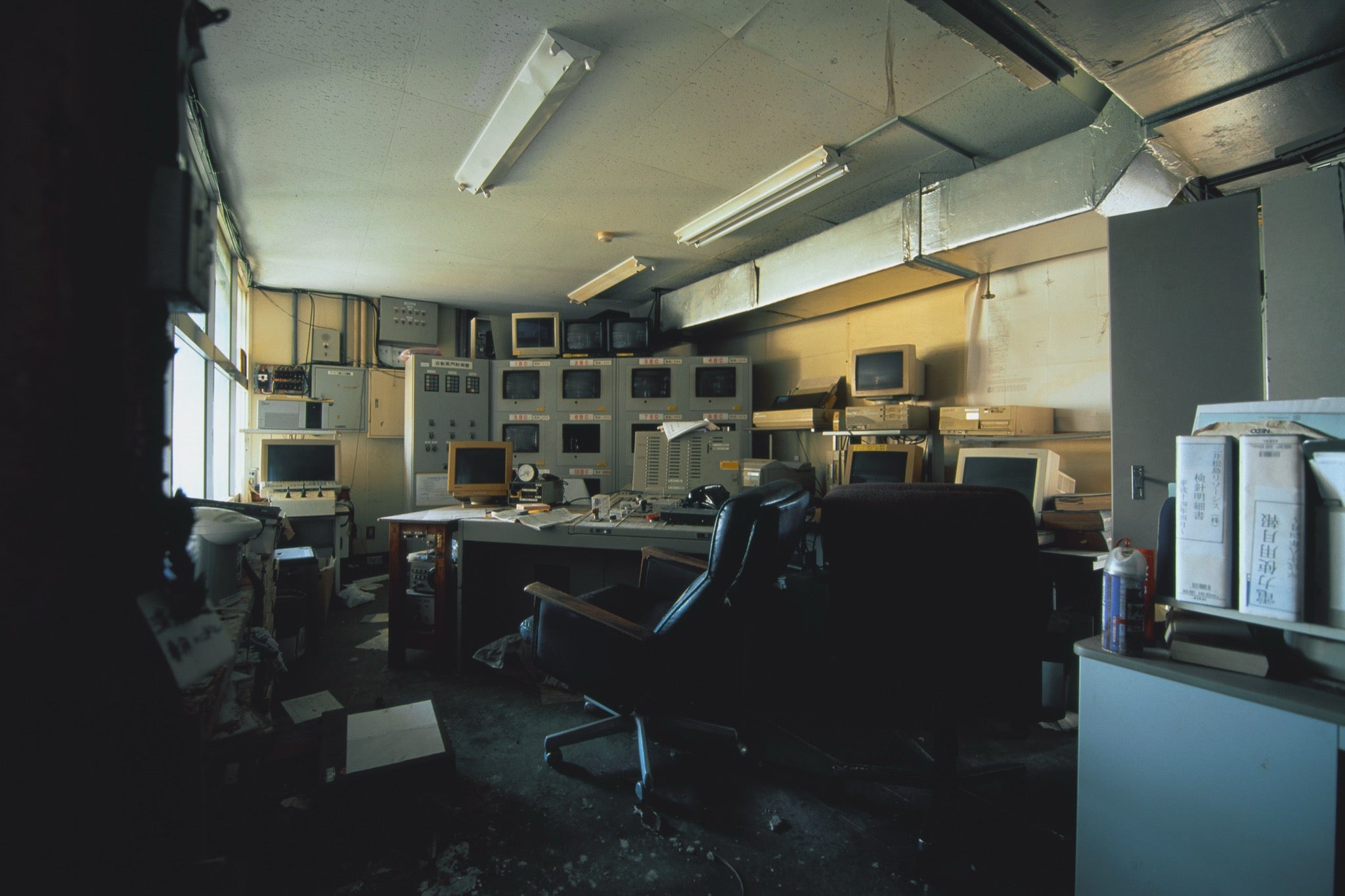 A bank of twelve dark CRT monitors set into and stacked above a central control console at the Ikeshima Coal Mine, with monthly report binders on a shelf to the right and daylight entering through a tall window at left.