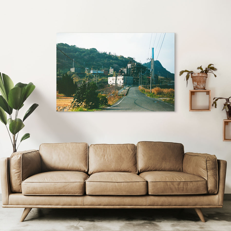 The chimney and boiler house of the coal-fired power station at Ikeshima, with an asphalt road curving past a small white-railed bridge in the foreground and a steep, densely vegetated hillside behind.