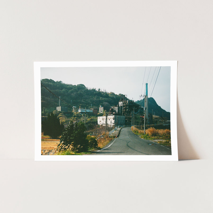 The chimney and boiler house of the coal-fired power station at Ikeshima, with an asphalt road curving past a small white-railed bridge in the foreground and a steep, densely vegetated hillside behind.