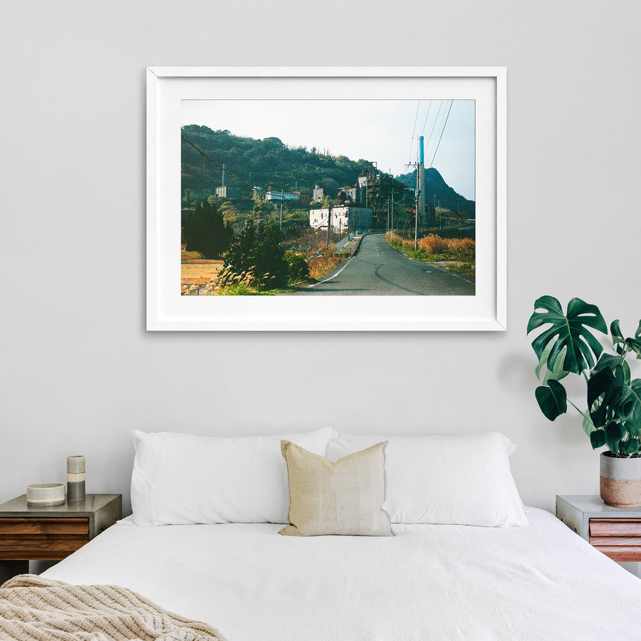The chimney and boiler house of the coal-fired power station at Ikeshima, with an asphalt road curving past a small white-railed bridge in the foreground and a steep, densely vegetated hillside behind.