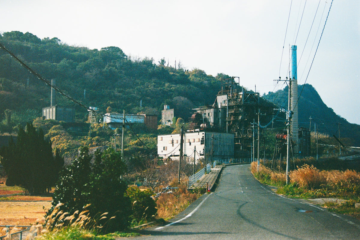 Road to the Ikeshima Coal-Fired Power Station, chimney and boiler house against a wooded hill with utility poles along the verge.