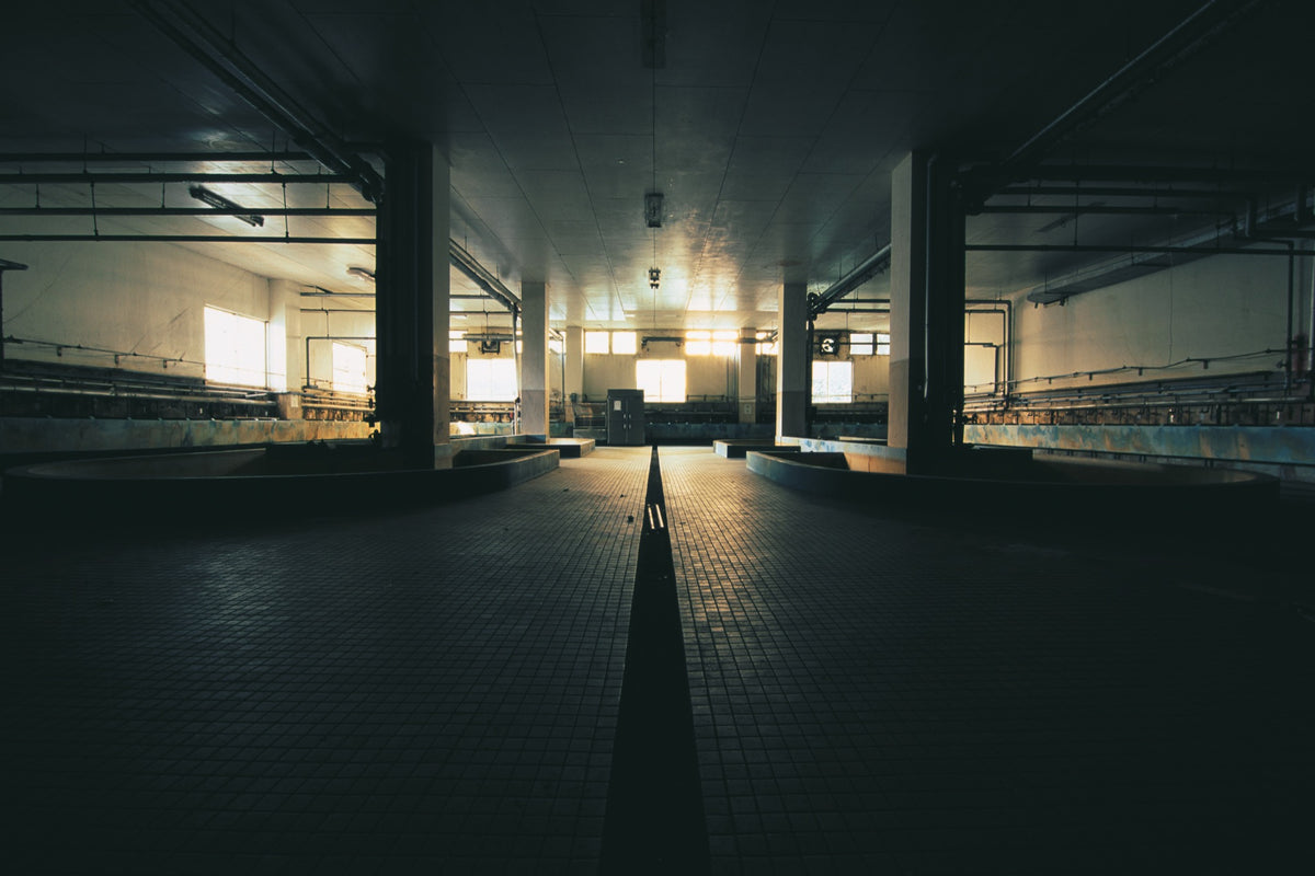 Interior of the communal bathhouse at Ikeshima Coal Mine, showing a central drainage channel between two U-shaped concrete baths, lit by golden light from square windows at the far end.