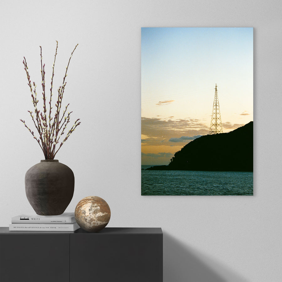 A lattice transmission tower silhouetted against a dusk sky on the forested ridgeline at Ikeshima, power lines extending from its upper arms toward calm water below.