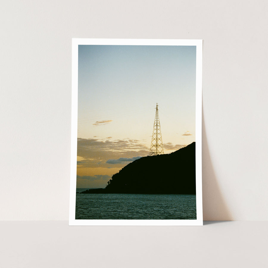 A lattice transmission tower silhouetted against a dusk sky on the forested ridgeline at Ikeshima, power lines extending from its upper arms toward calm water below.