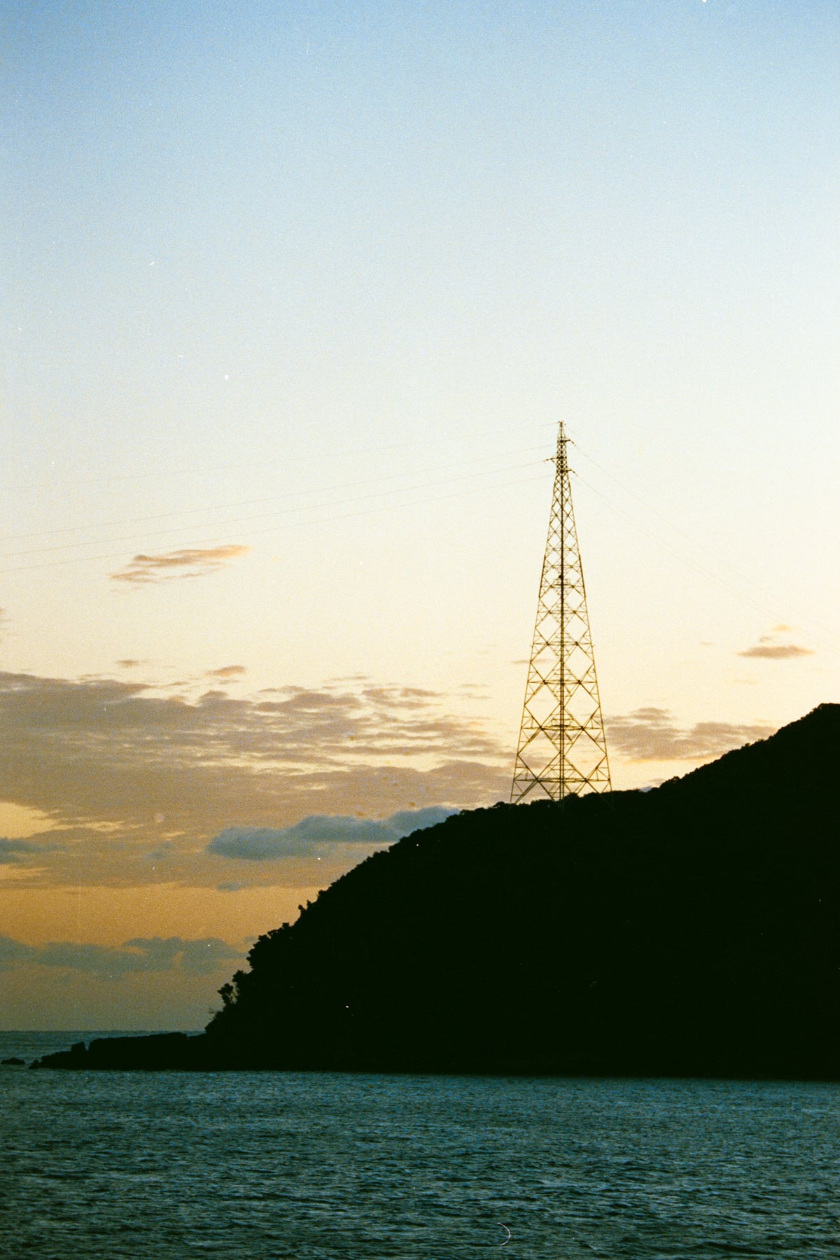 A lattice transmission tower silhouetted against a dusk sky on the forested ridgeline at Ikeshima, power lines extending from its upper arms toward calm water below.