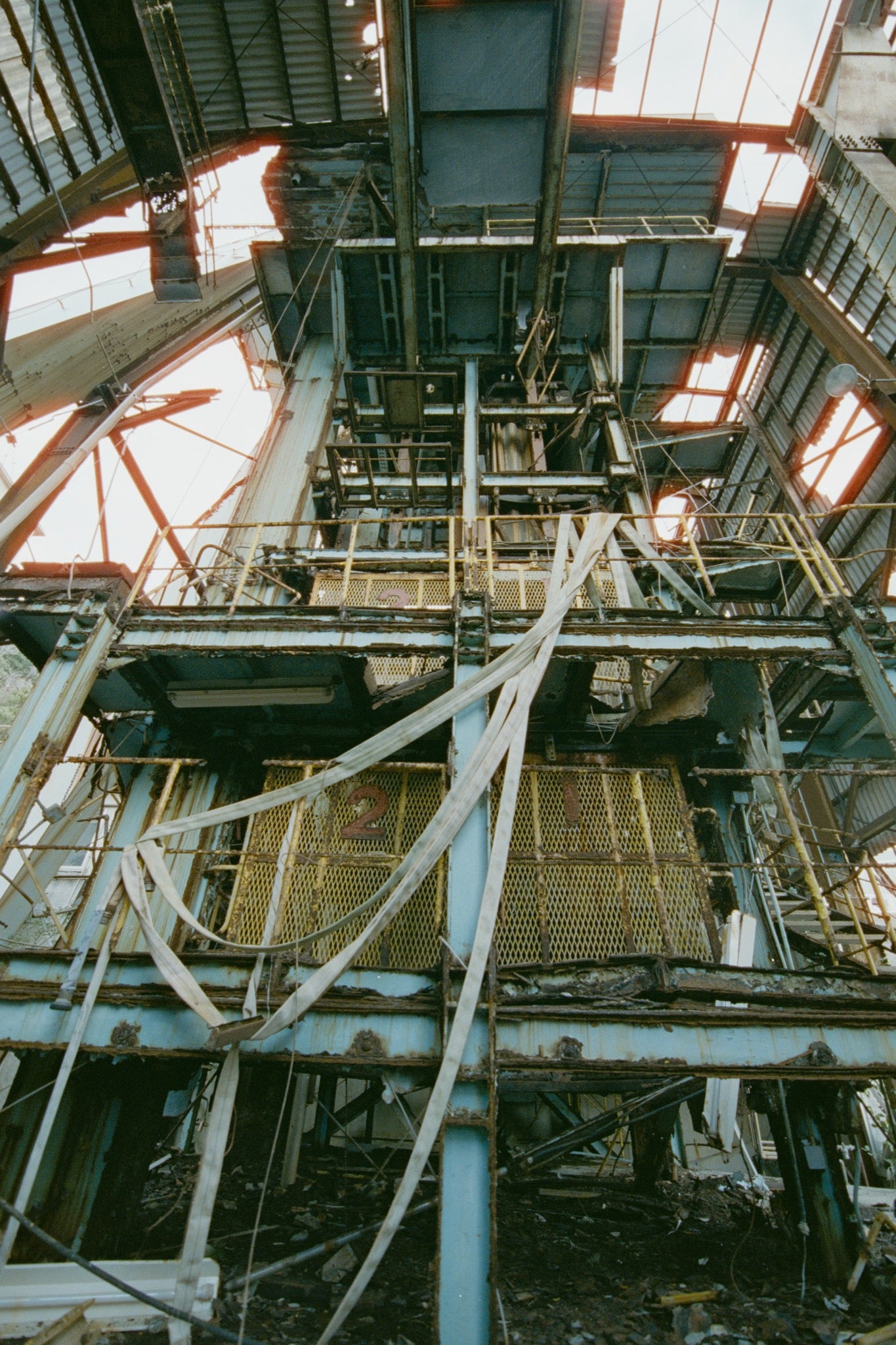 Corroded steel framing and two numbered yellow mesh barrier panels inside the main mining building at Ikeshima Coal Mine, with white fabric hanging loosely across them and daylight entering through holes in the corrugated roof above.