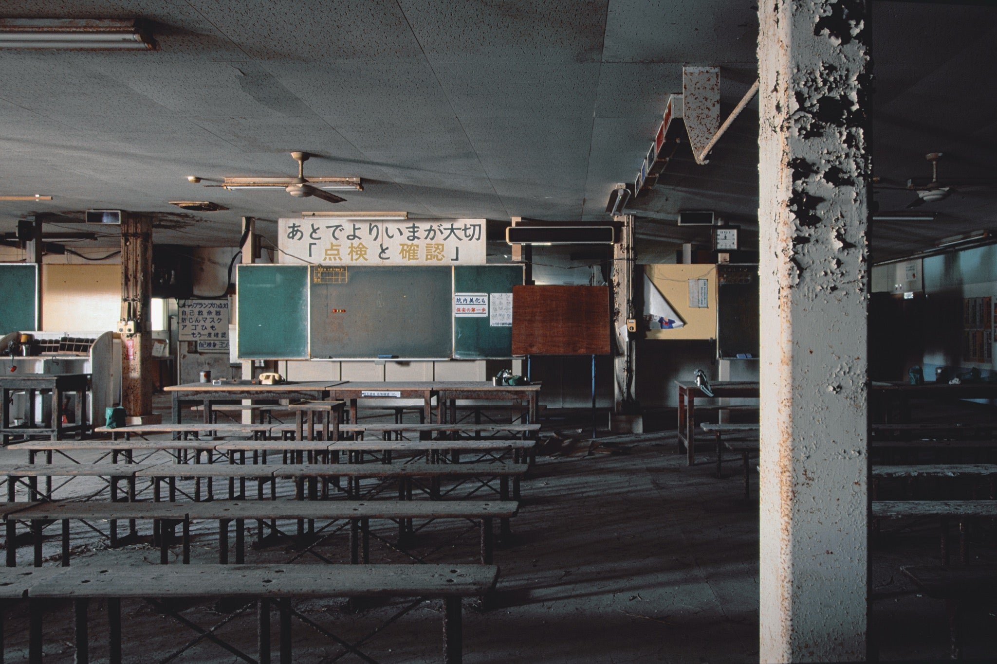 A dust-covered briefing room at Ikeshima Coal Mine, with rows of wooden tables and bench seating facing two dark green chalkboards, a rotary telephone on a table, and a safety sign on the rear wall carrying Japanese text.