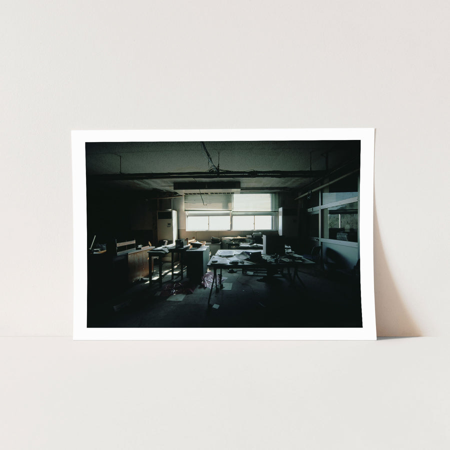 Interior of a derelict office at Ikeshima Coal Mine, with horizontal blinds casting banded light across paper-strewn desks, a large dark monitor, a white air conditioning unit, and a sagging ceiling with visible water damage.