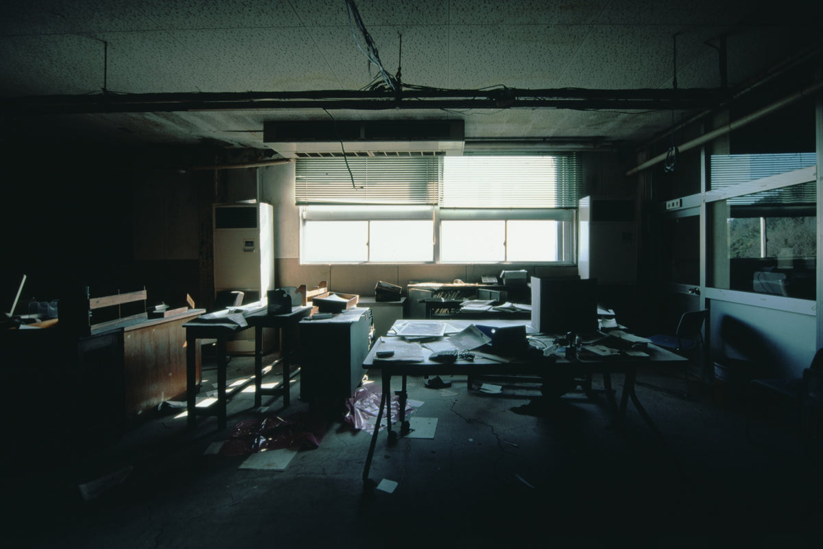 Interior of a derelict office at Ikeshima Coal Mine, with horizontal blinds casting banded light across paper-strewn desks, a large dark monitor, a white air conditioning unit, and a sagging ceiling with visible water damage.