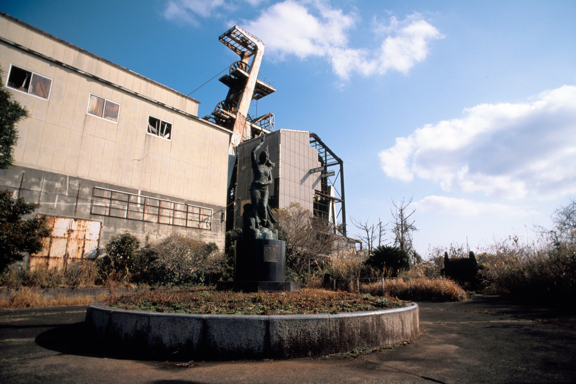 A bronze figure with one arm raised on a concrete plinth, with the rusted steel headframe and winding house of Ikeshima Coal Mine visible behind it.