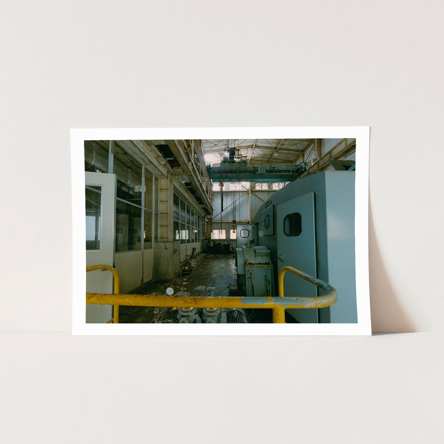 Interior of the winding house at Ikeshima Coal Mine, showing a light-blue winding engine with panelled doors, a teal overhead gantry crane, glass-paned windows, and a pressure gauge on the concrete floor.