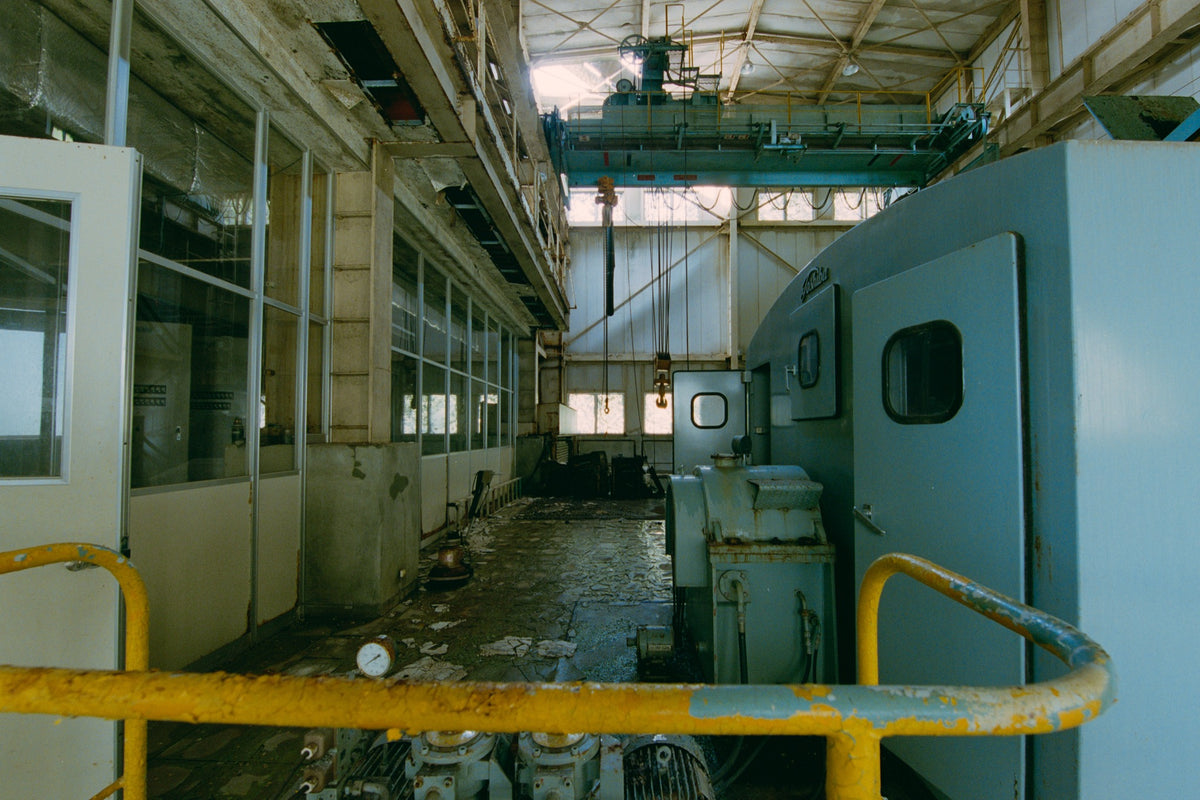 Interior of the winding house at Ikeshima Coal Mine, showing a light-blue winding engine with panelled doors, a teal overhead gantry crane, glass-paned windows, and a pressure gauge on the concrete floor.