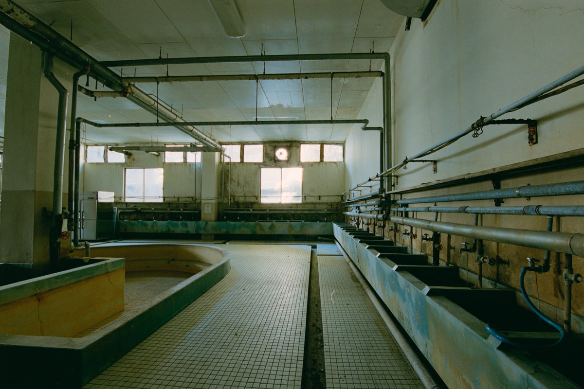 Corroded rectangular basins with individual taps along the wall of the miners' wash house at Ikeshima Coal Mine, with a curved yellow-and-green trough in the foreground above white floor tiles.
