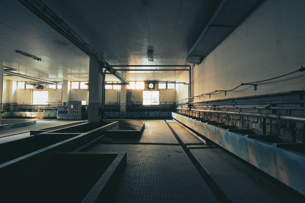 Interior of the miners' bathhouse at Ikeshima Coal Mine, with a clock above far windows, concrete washing troughs across the tiled floor, and a rusted spigot trough along the right wall.