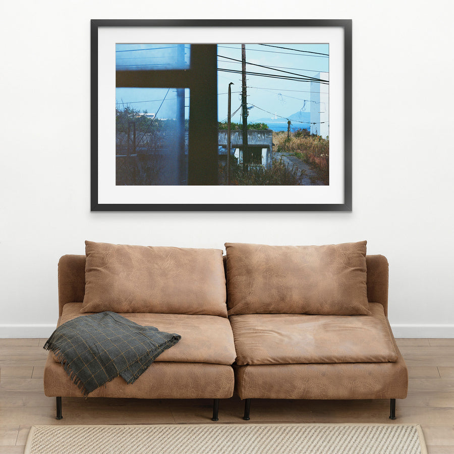 View through a dark window frame at Ikeshima showing two utility poles with tangled wires against pale sky, dry grass, a concrete structure, and a white lighthouse on a distant landmass across open water.