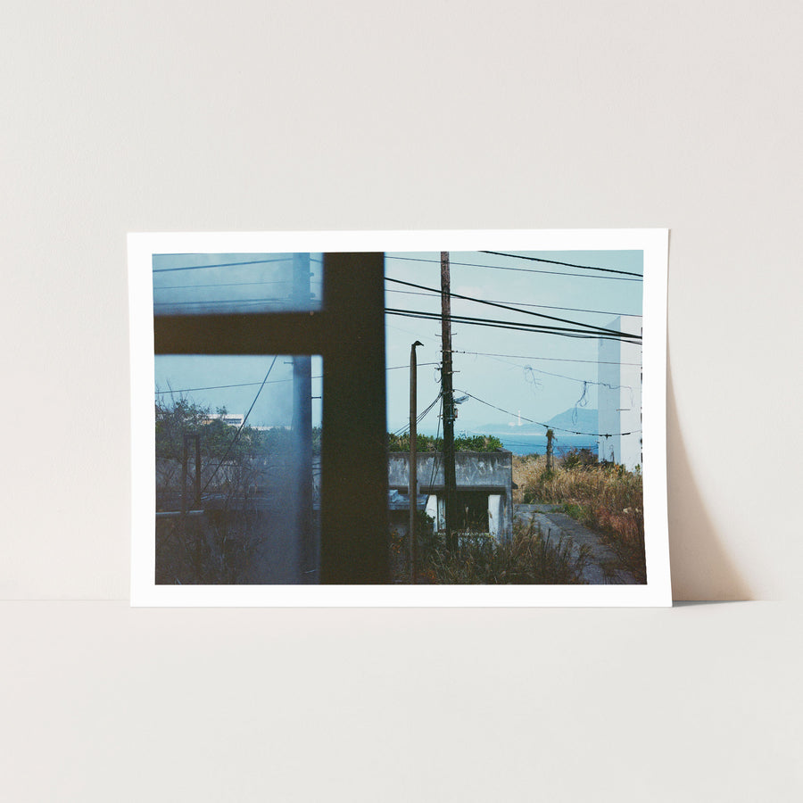 View through a dark window frame at Ikeshima showing two utility poles with tangled wires against pale sky, dry grass, a concrete structure, and a white lighthouse on a distant landmass across open water.