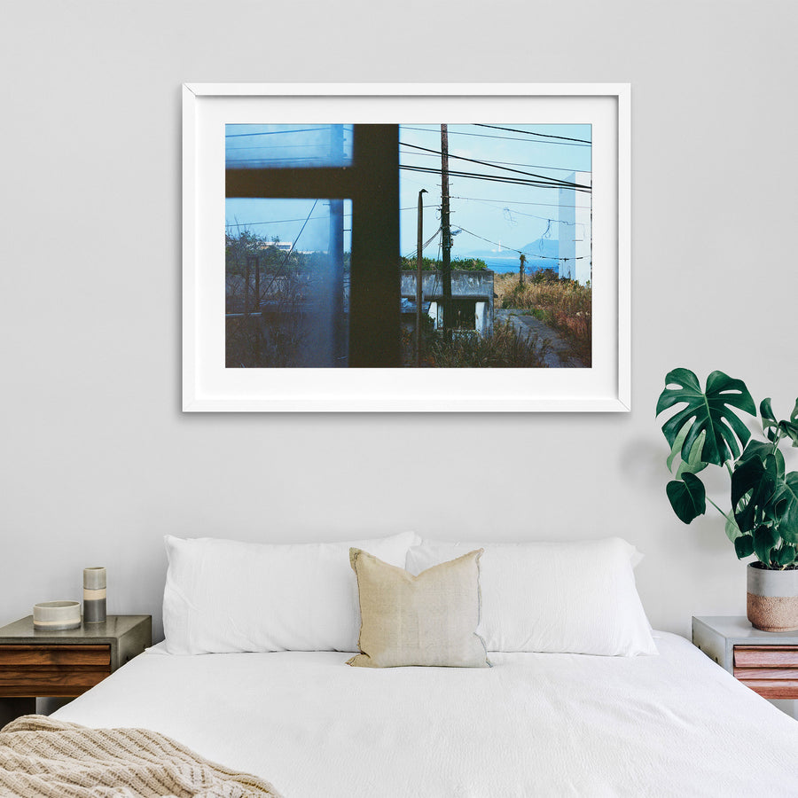 View through a dark window frame at Ikeshima showing two utility poles with tangled wires against pale sky, dry grass, a concrete structure, and a white lighthouse on a distant landmass across open water.