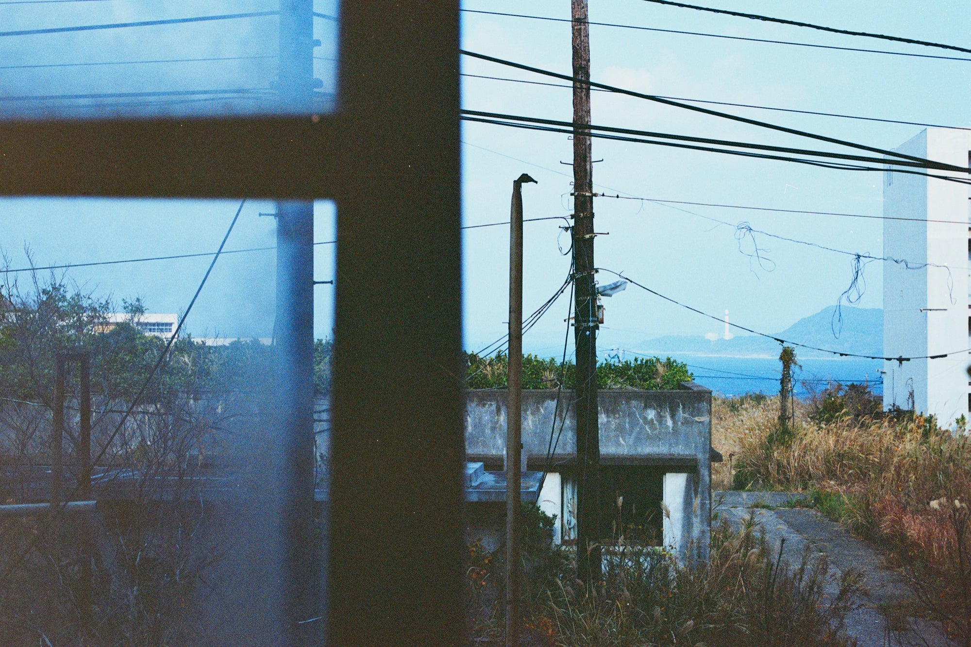 View through a dark window frame at Ikeshima showing two utility poles with tangled wires against pale sky, dry grass, a concrete structure, and a white lighthouse on a distant landmass across open water.