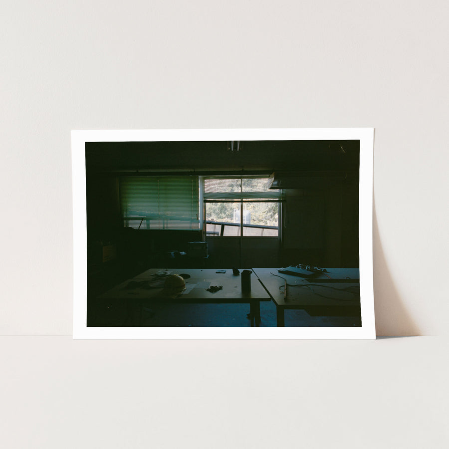 A yellow hard hat and scattered tools on a cluttered workbench inside the workshops and safety offices at Ikeshima Coal Mine, with buckled blinds and daylight through a glass window behind.