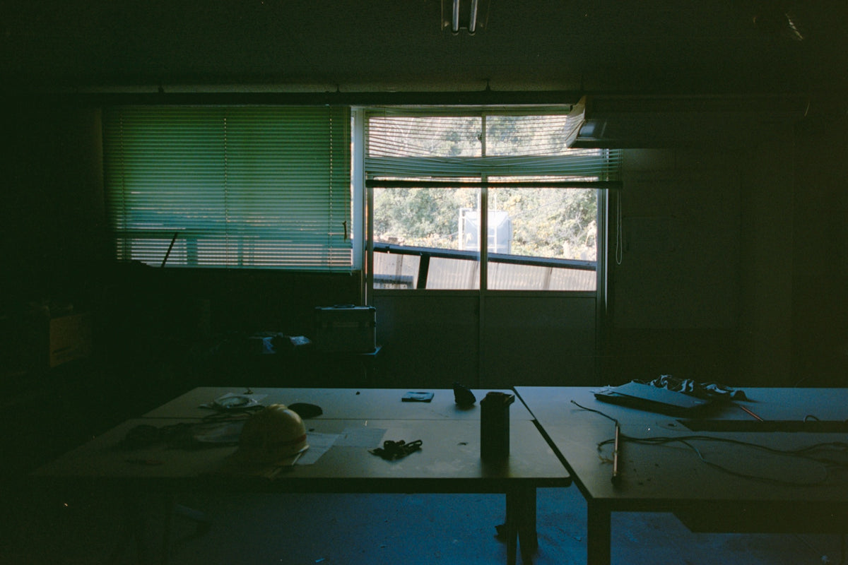 A yellow hard hat and scattered tools on a cluttered workbench inside the workshops and safety offices at Ikeshima Coal Mine, with buckled blinds and daylight through a glass window behind.