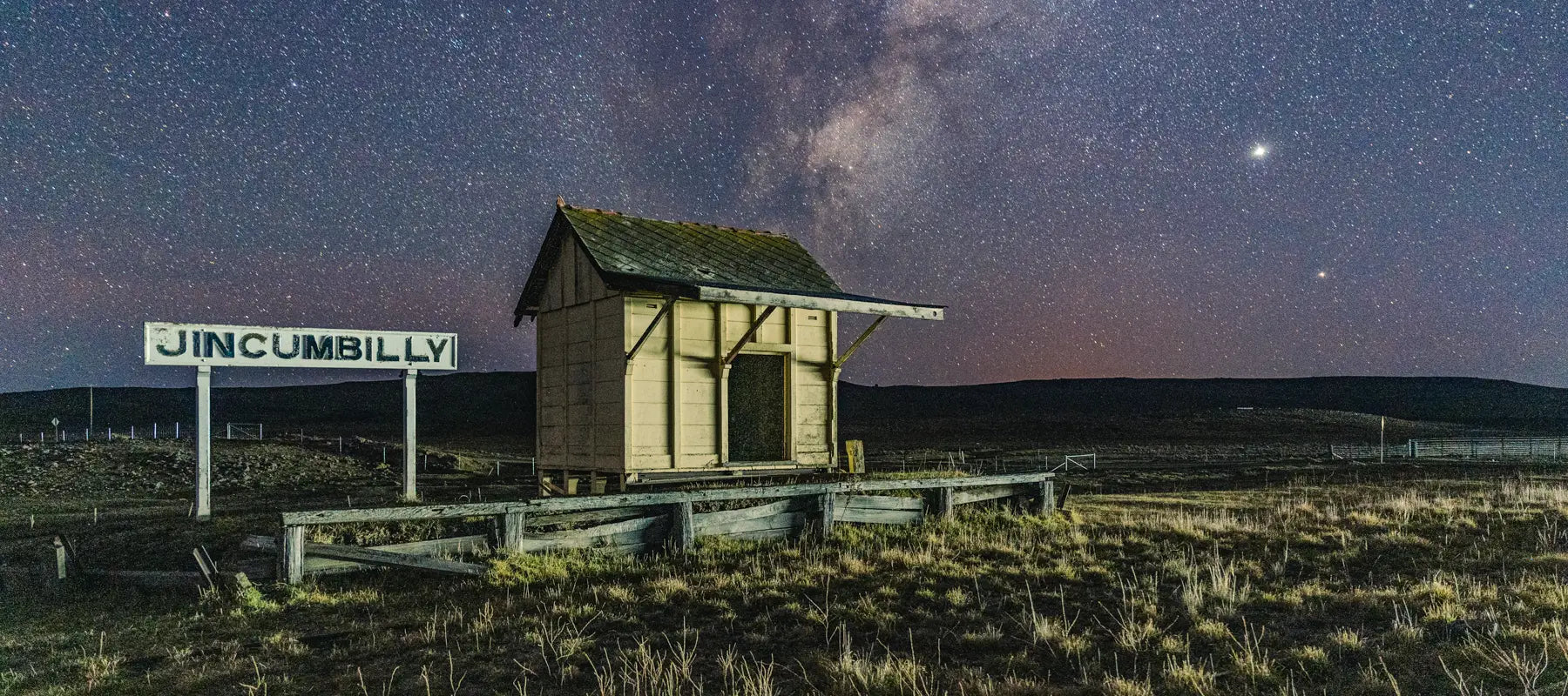 Abandoned Jincumbilly Railway Station under a star-filled night sky. The weathered wooden platform and faded station sign stand in contrast to the vast open landscape, illuminated by the Milky Way overhead