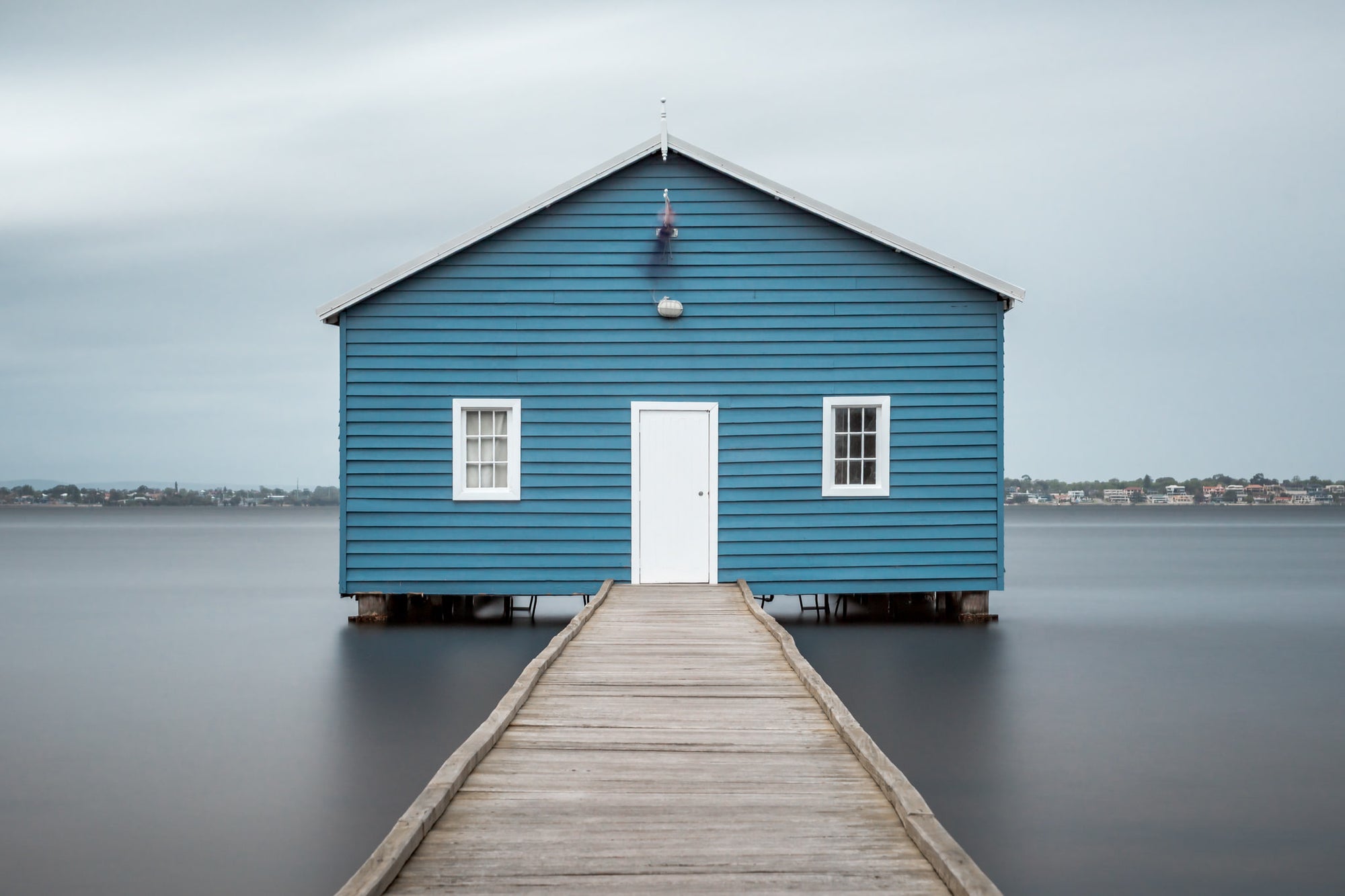 Crawley Edge Boatshed at Landscapes, a single blue timber boatshed sits on steel piles over flat grey water.