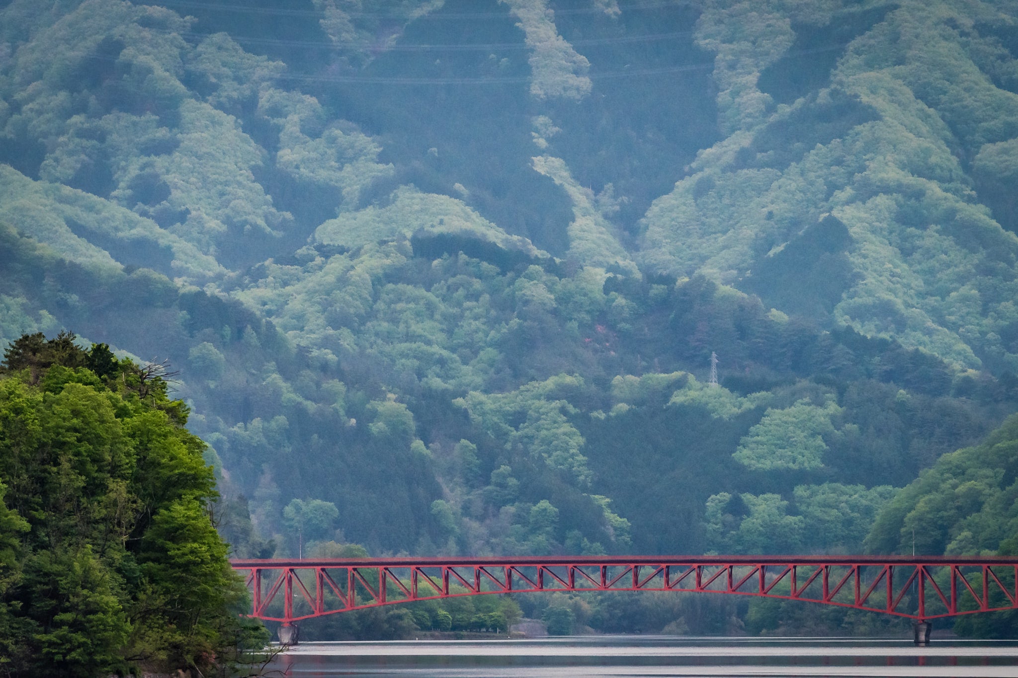 Kusaki Dam Lake at Landscapes, a red steel bridge dwarfed by mountains stretches over a lake.