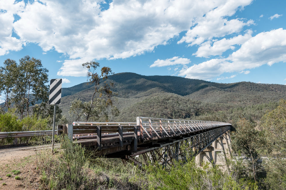 View along the road approach to McKillops Bridge in Deddick Valley, with a black and white chevron sign at left and the single-lane timber-decked steel truss bridge spanning the Snowy River, forested hills rising beyond under an overcast sky.