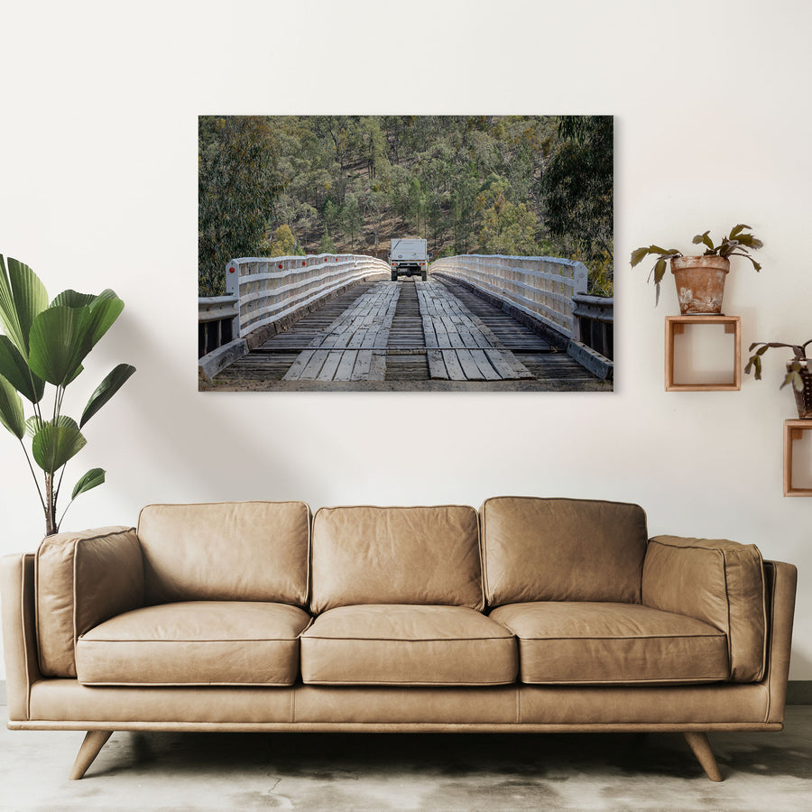 A white vehicle towing a camper crosses McKillops Bridge head-on along the timber running planks, tail-lights lit, between tall white handrails with a forested hillside behind.