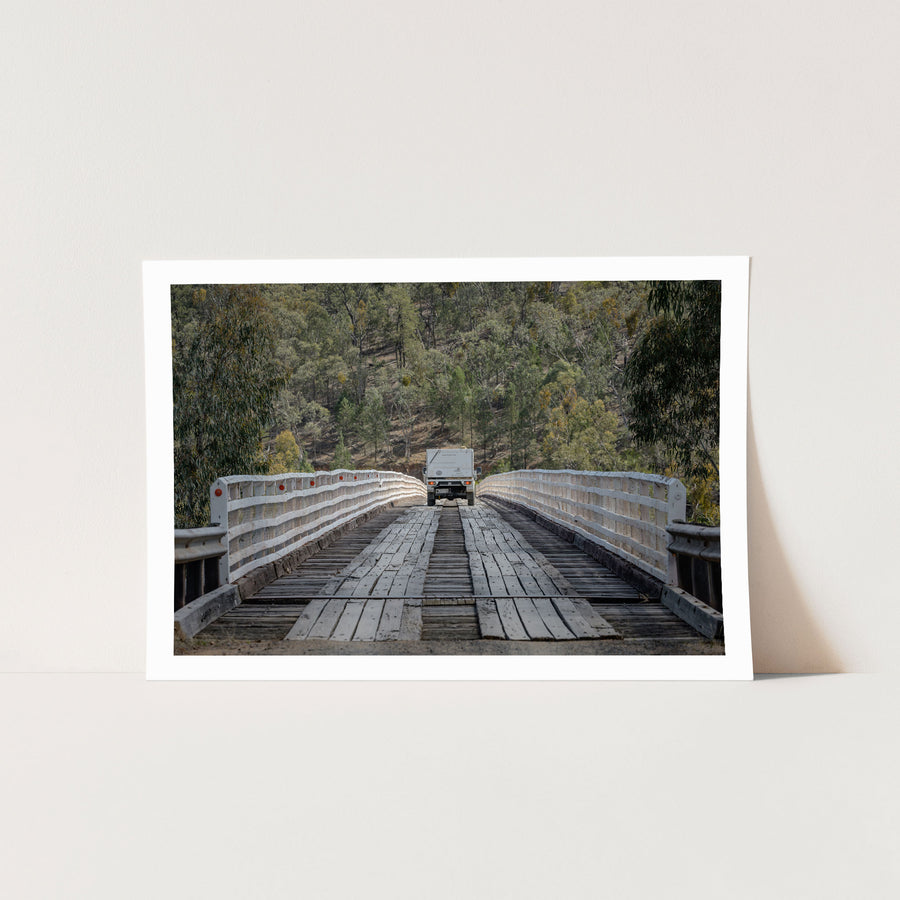 A white vehicle towing a camper crosses McKillops Bridge head-on along the timber running planks, tail-lights lit, between tall white handrails with a forested hillside behind.