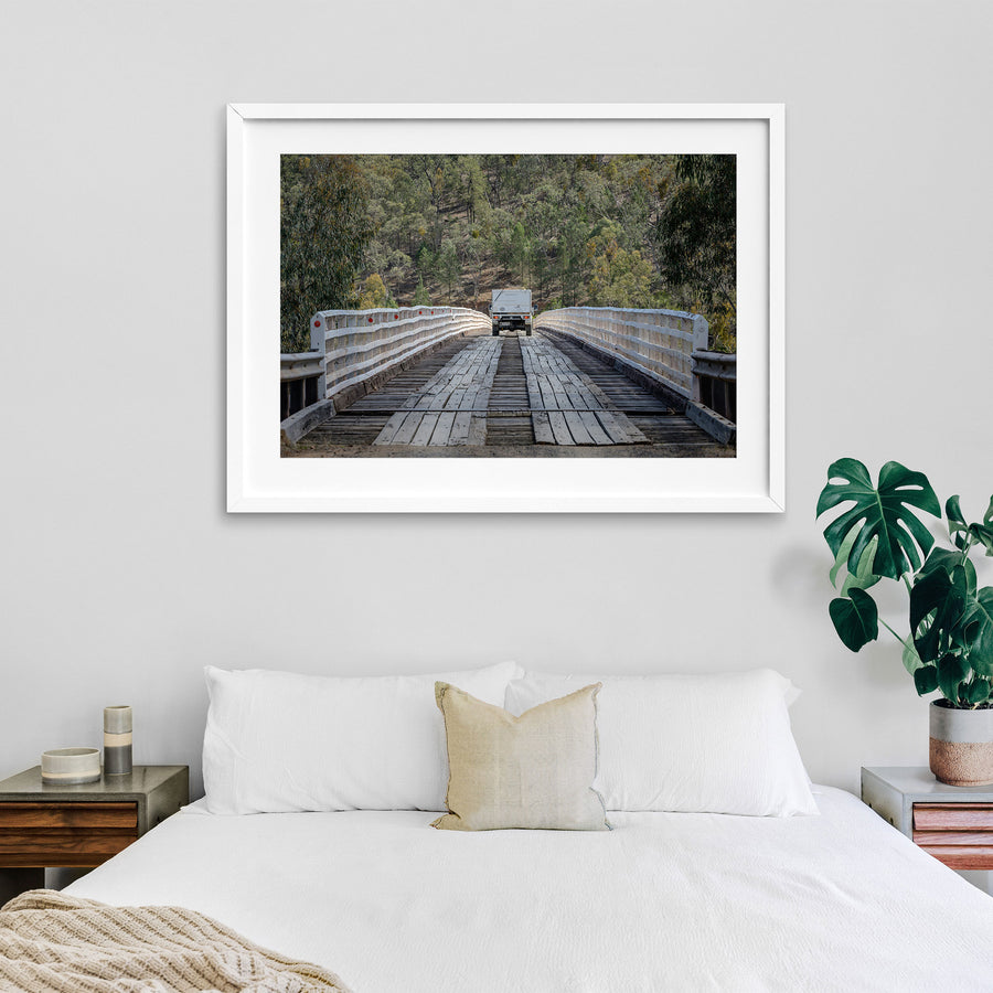A white vehicle towing a camper crosses McKillops Bridge head-on along the timber running planks, tail-lights lit, between tall white handrails with a forested hillside behind.