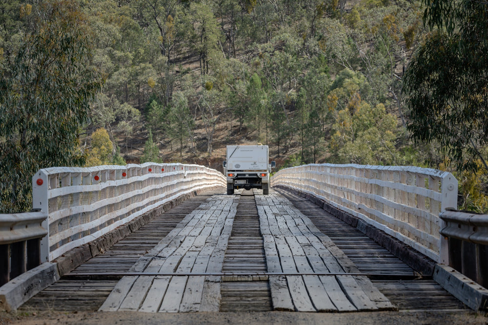 A white vehicle towing a camper crosses McKillops Bridge head-on along the timber running planks, tail-lights lit, between tall white handrails with a forested hillside behind.
