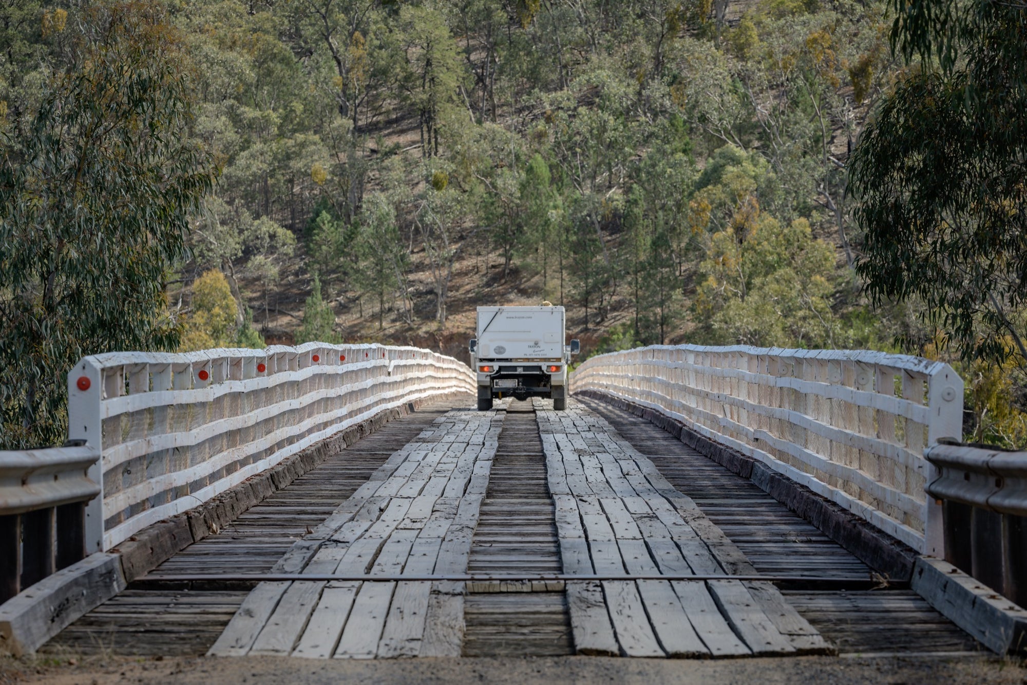 A white vehicle towing a camper crosses McKillops Bridge head-on along the timber running planks, tail-lights lit, between tall white handrails with a forested hillside behind.