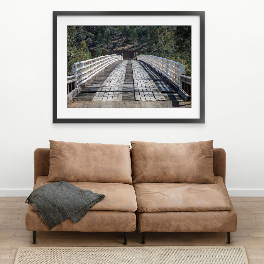 End-on view down the worn timber deck of McKillops Bridge in Deddick Valley, white handrails with red reflectors flanking the single lane as it runs toward a wooded hillside cutting.